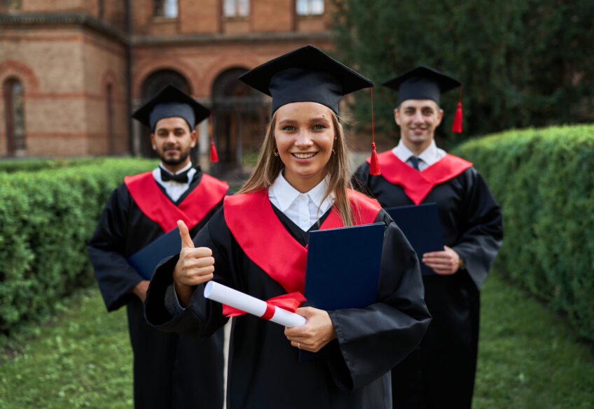 Graduate female shows like with her friends in graduation gowns holding diploma and smiling at camera.